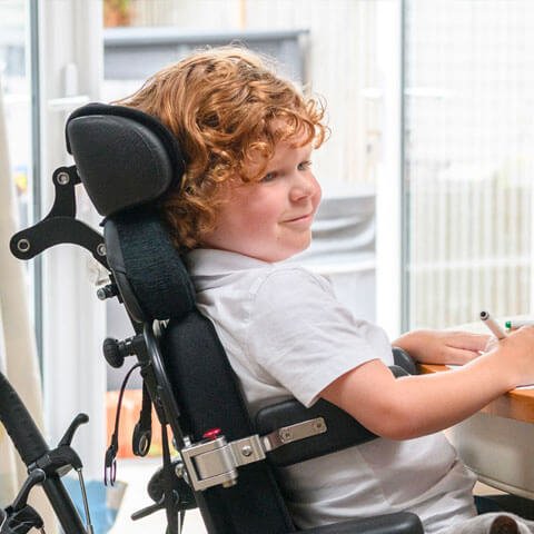 Young boy in a wheelchair sitting at a desk smiling and writing