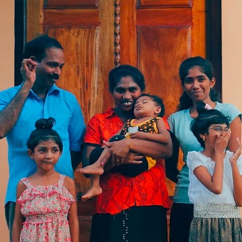 Sri Lankan family stood outside a family. A mother holding a baby next to her husband and three children